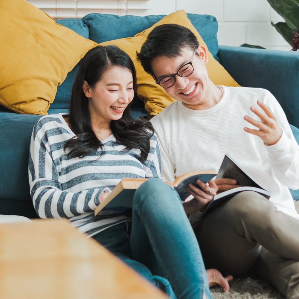 A couple is sitting on the floor in front of a couch, looking at books and laughing.