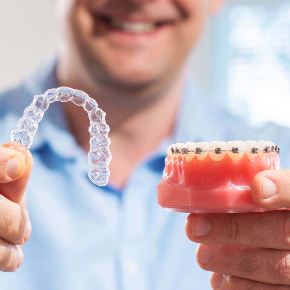 A dentist holds up an example of a mouth with Invisalign clear aligners in, and a set of teeth with traditional braces.