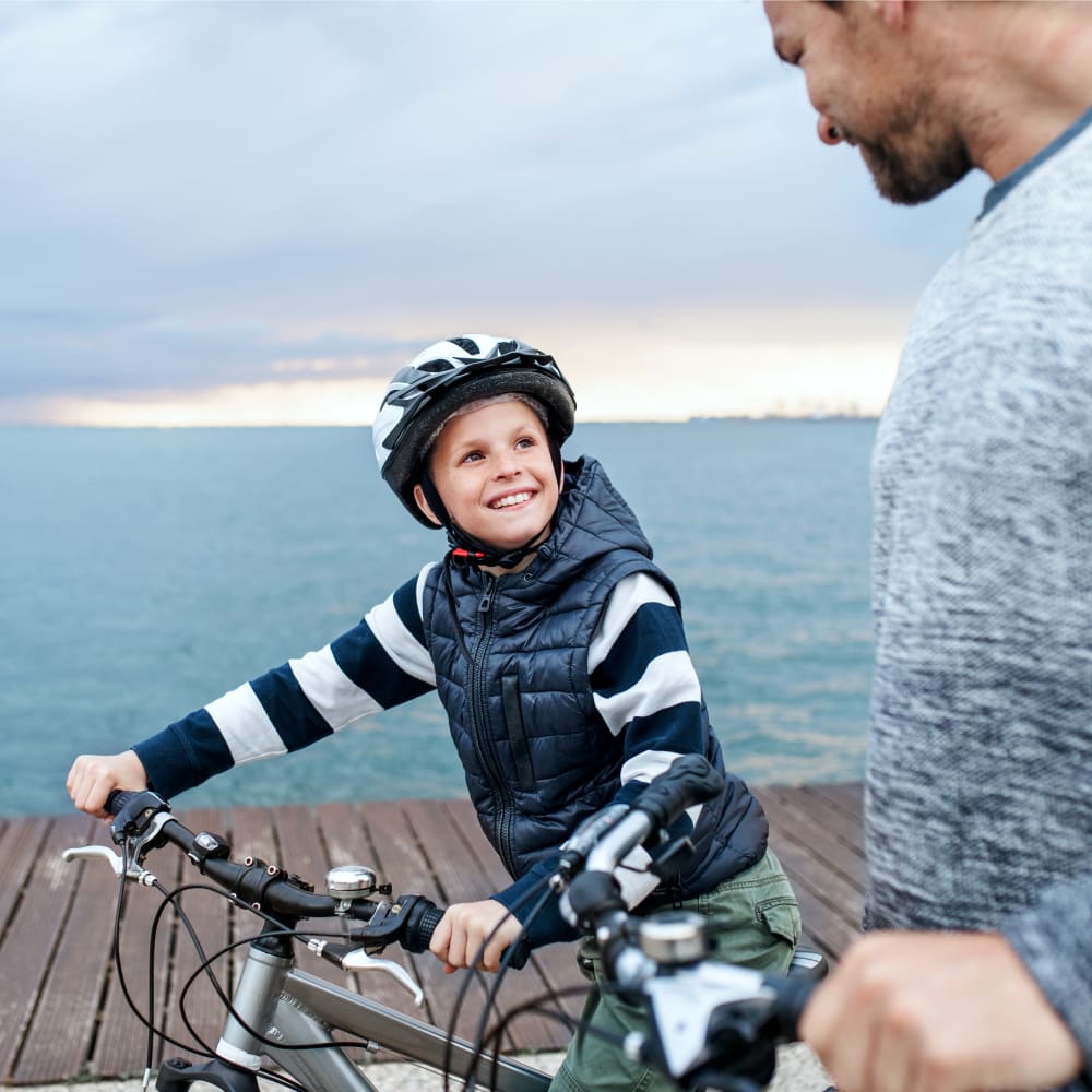 A young boy is riding his bike down a boardwalk smiling. An adult man is beside him.