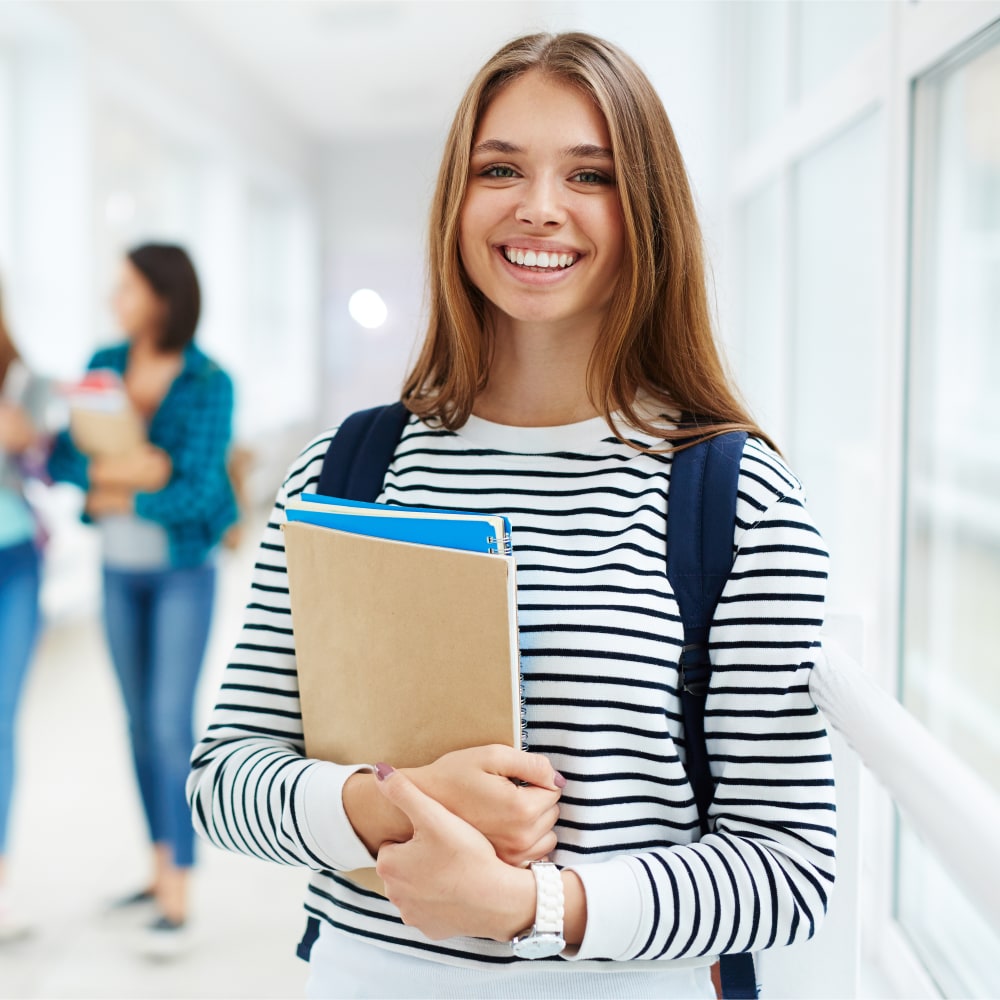 A teenage girl is smiling. She is holding books and wearing a backpack.